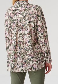 Blouse à motifs floraux en tissu léger, avec une base pastel ornée de fleurs vertes, roses et bleues. Manches longues avec des poignets à boutons.