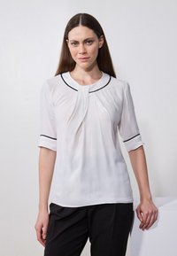 Blouse blanche à manches courtes et décolleté rond, ornée de piping noir le long du col et des manches, avec un design plissé à l'avant.