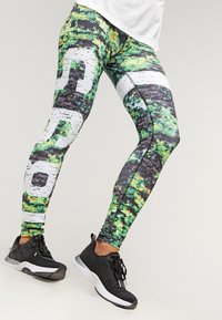 Vícebarevné legíny se zeleným, černým a bílým abstraktním vzorem, s výrazným bílým logem, vyrobené z elastického a přiléhavého materiálu.