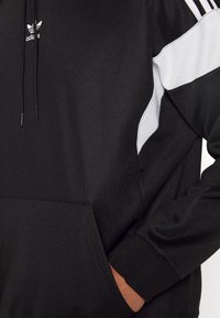Black hoodie with white accents, adjustable drawstring, front pocket, and three white stripes on the shoulder, made of textured fabric.