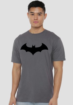 Young man wearing a gray t-shirt with a large black bat symbol on the chest, standing against a plain light background.