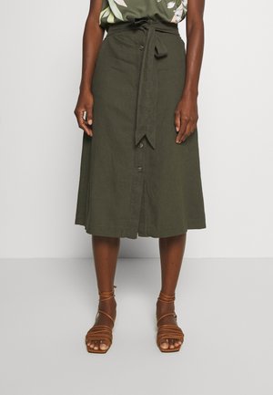 Jupe midi boutonnée devant de couleur vert foncé avec ceinture nouée à la taille, portée avec des sandales marron à lanières sur une personne debout.