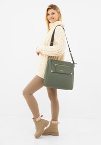 Borsa in finta pelle verde con tasca frontale con cerniera, dettagli in hardware dorato e tracolla regolabile. Il modello indossa un maglione crema e leggings marroni.