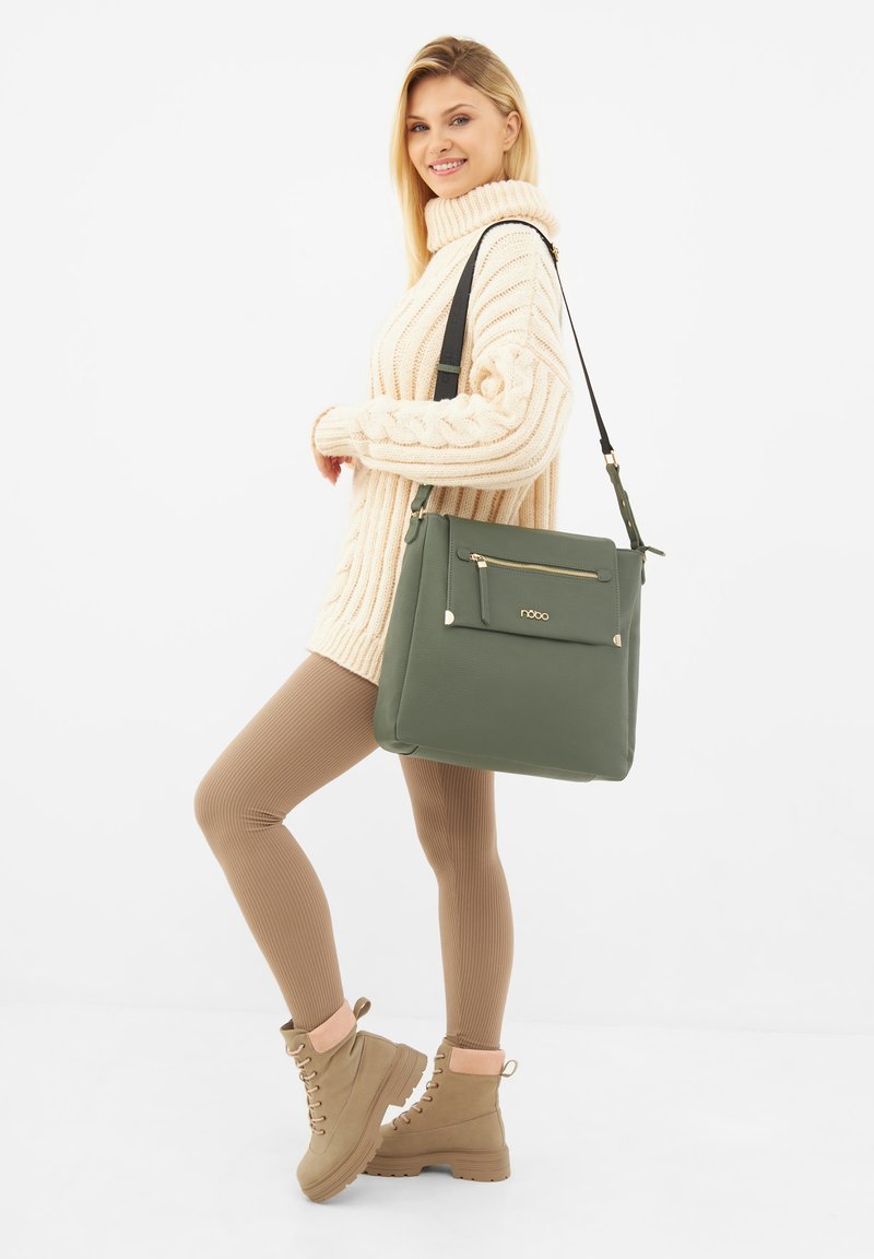 Borsa in finta pelle verde con tasca frontale con cerniera, dettagli in hardware dorato e tracolla regolabile. Il modello indossa un maglione crema e leggings marroni.