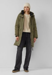 Olijfgroene parka jas met een capuchon, twee voorzakken, zwarte voering, gecombineerd met een crème gebreide trui en grijze jeans. Zwarte schoenen.
