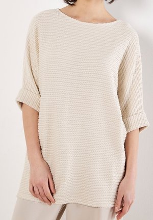 Vrouw draagt een losse, off-white textuurtrui met opgerolde mouwen tot aan de elleboog en lichtgekleurde broek.