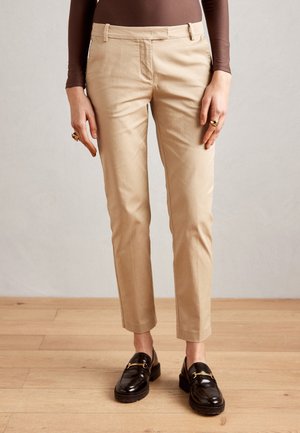 Person trägt beige, schmal geschnittene Hose und schwarze Leder-Loafers, steht auf Holzboden vor schlichter Wand.