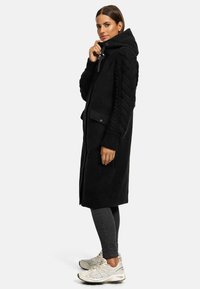 Black long coat with a hood, textured knit sleeves, and side pockets, paired with dark jeans and light sneakers.