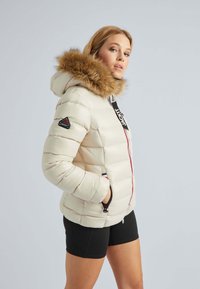 Crèmekleurige pufferjack met een bontgevoerd capuchon, contrasterende rits en zijzakken. Bevat een merkembleem op de mouw en een geborduurde textuur.