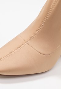 Bottine beige à bout pointu, en tissu lisse avec des coutures visibles, présentant une coupe ajustée et une silhouette élégante.