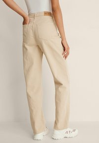 Pantalones de tiro alto beige con corte recto, con bolsillos traseros y una cinturilla con marca. Tejido de algodón texturizado y costuras mínimas.