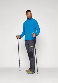 Top azul de manga comprida com fecho de um quarto, calças cinzentas com detalhes pretos e verdes, bastões de caminhada e sapatos de caminhada esverdeados com solas azuis.