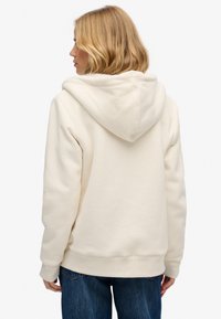 Sweatshirt couleur crème avec une grande capuche douce, des poignets et un ourlet côtelés. Le tissu semble moelleux et lisse, avec une coupe décontractée.