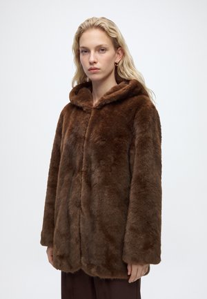 Femme aux cheveux clairs portant un manteau à capuche en fausse fourrure marron, debout devant un fond clair uni avec une expression neutre.