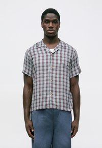Uomo con camicia a maniche corte a quadri in rosso e bianco, abbinata a pantaloni blu, in piedi contro uno sfondo chiaro e uniforme.