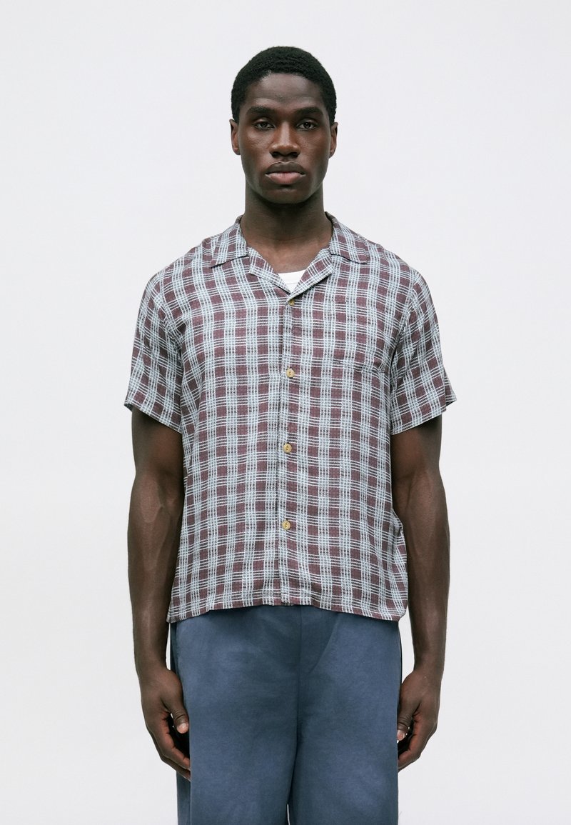 Uomo con camicia a maniche corte a quadri in rosso e bianco, abbinata a pantaloni blu, in piedi contro uno sfondo chiaro e uniforme.