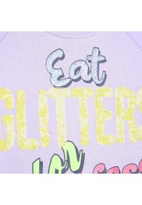 Fioletowa bluza z przędzy z wielokolorowym tekstem z cekinów. "Jedz" w iryzującym niebieskim, "BROKATY" w żółtych cekinach, z zielonymi akcentami poniżej.
