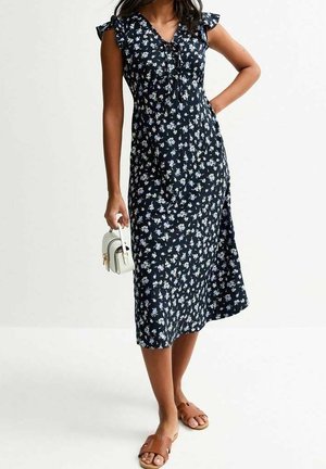 Navy floral dress with short ruffle sleeves, a tie detail at the neckline, and a flowing silhouette. Paired with a small white handbag and sandals.