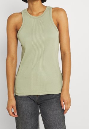 Kobieta w jasnozielonym prążkowanym topie bez rękawów i ciemnoszarych dżinsach, stojąca na białym tle.