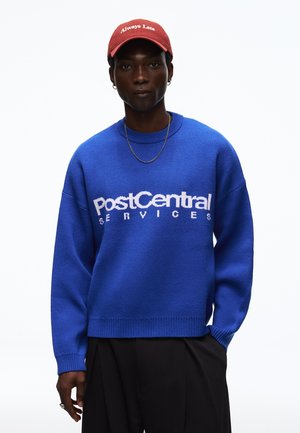 Mann mit roter Kappe mit dem Text "Always Late" und blauem Pullover mit dem Text "PostCentral Services", stehend vor weißem Hintergrund.