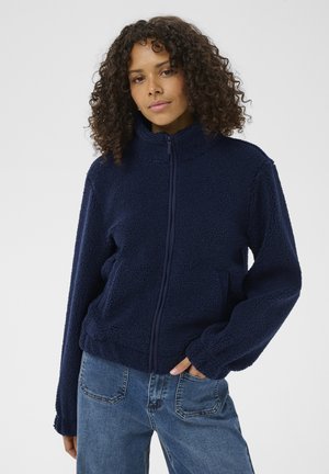 Mladá žena s kučeravými vlasmi, oblečená v námorníckom modrom fleecovom pulóvri so zipsom a modrých džínsoch, stojaca s jednou rukou v vrecku.