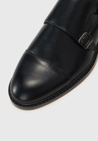 Black leather shoe featuring a pointed toe design, single strap with a metal buckle, and subtle stitching details around the edges.