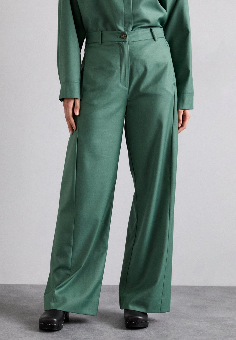 Pantaloni larghi verdi realizzati in un tessuto morbido, con chiusura frontale con bottone, passanti per cintura e tasche laterali. Zoccoli neri completano il look.
