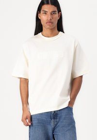 Off-white bomull T-skjorte med korte ermer, med en hevet "GUESS" logo foran. Kombinert med blå denim jeans.
