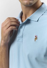 Svijetloplava polo majica s ovratnikom, s malim izvezenim logotipom na prsima i dva gumba na preklopu.