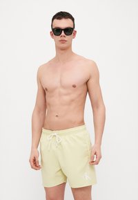 Shorts de bain jaune clair avec taille élastique et cordon de serrage ; présente un logo "ck" blanc sur le côté gauche. Modèle portant des lunettes de soleil noires.