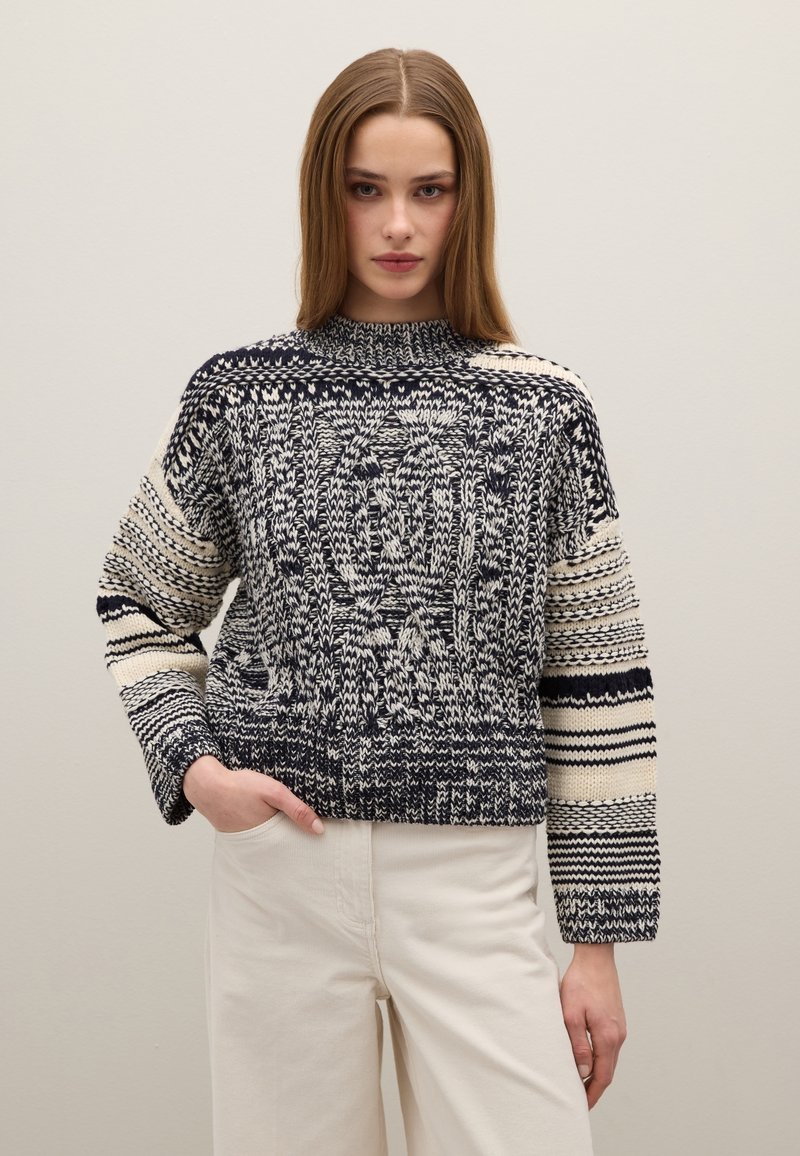 Gestrickter Pullover mit hohem Kragen, versehen mit marineblauen und cremefarbenen Mustern, voluminösen Ärmeln und einer strukturierten Oberfläche. Kombiniert mit hellen Hosen.