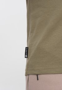 Olivgrünes Baumwoll-T-Shirt mit tailliertem Schnitt, versehen mit einem schwarzen Logo-Tag an der Seite und einer glatten Haptik.