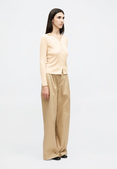 Γυναίκα που στέκεται, φοράει μπεζ ριγωτή ζακέτα με κουμπιά και χακί παντελόνια ψηλής μέσης με φαρδιά μπατζάκια, μαζί με μαύρα παπούτσια.