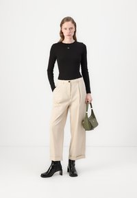 Top nero a maniche lunghe, pantaloni beige a vita alta e gamba larga, stivaletti neri alla caviglia e una piccola borsa verde. Design minimalista, texture lisce.
