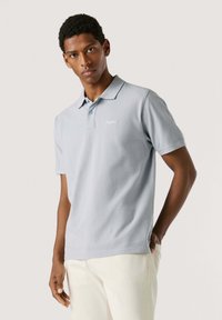 Polo gris clair à manches courtes, à patte de boutonnage à deux boutons, avec logo brodé sur la poitrine gauche. Texture lisse, matière en coton.