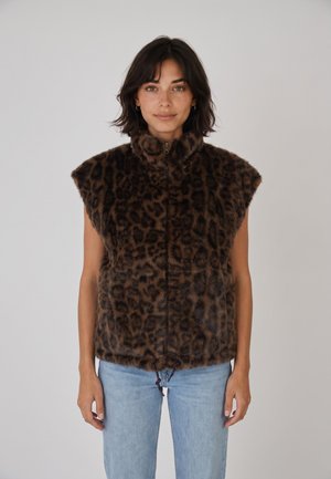 Femme aux cheveux courts et foncés porte un gilet en fourrure sans manches imprimé léopard marron et noir et un jean bleu clair, debout sur un fond uni.