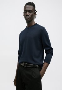 Tumesinist kudumitööd kampsun ümara kaelusega ja pikkade varrukatega, mille rinnal väike logo. Sobitatud mustade pükstega.