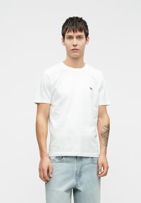 White cotton t-shirt with short sleeves, featuring a small blue embroidered logo on the left chest. Smooth texture, classic fit.