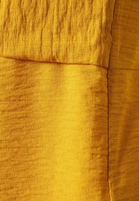 Texturovaná tkanina v bohatém žlutém odstínu, s krabatým povrchem a různými vzory a hladkými částmi. Žádné viditelné akcenty ani kovové prvky.