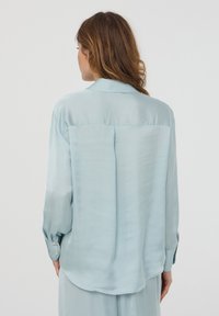 Blusa in satin azzurro chiaro con una vestibilità comoda, maniche lunghe e polsini con bottone. Presenta un yoke posteriore e un orlo arrotondato. Texture liscia.