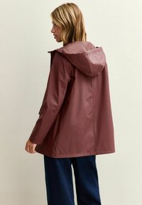 Röd-burgundy vattentät jacka med huva, med rak passform, långa ärmar och sidofickor, sedd bakifrån.