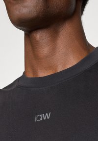 Čierne priliehavé tričko s okrúhlym výstrihom, s malým sivým logom "ICIW" na hrudi. Hladká textúra látky.