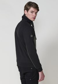 Sudadera negra con cremallera y cuello alto, que presenta un panel texturizado gris, cordón ajustable y un parche con el logo en la manga izquierda.