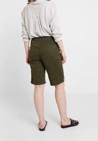 Shorts cargo verts olive avec une coupe droite et des poches arrières, associés à une chemise à manches longues à carreaux légers et des sandales noires à enfiler.