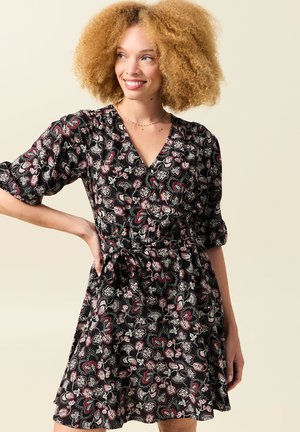 Femme aux cheveux blonds bouclés portant une robe portefeuille noire à fleurs, souriante et regardant sur le côté devant un fond neutre.