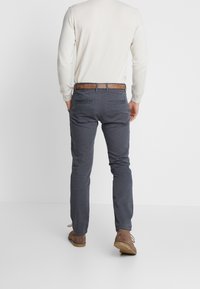 Chinos da uomo grigio scuro con una vestibilità slim, dotati di cinque tasche, una cintura marrone e una texture sottile. Indossati con una maglietta a maniche lunghe color crema chiaro.