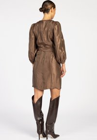 Femme debout de dos, portant une robe brune brillante avec des manches bouffantes et des bottes à talons haut, marron foncé, jusqu'aux genoux.