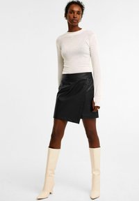 Sort læderomslagsskørt med en asymmetrisk nederdel, parret med en hvid langærmet top. Hvide knæhøje støvler med hæl fuldender outfittet.