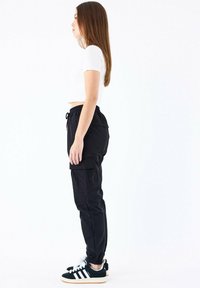Sorte cargo bukser med sidelommer, elastisk talje og snoretræk. Parret med sort-hvide sneakers. Modellen ses fra siden.