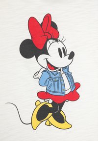 Grafiska t-krekls ar karikatūru varoni, kas ģērbjas zilo džinsa jaku, sarkanu kleitu un dzeltenām kurpēm, uz gaiša auduma fona.